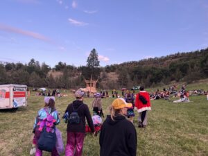 Festival attendees in colorful costumes walking across an open field toward a wooden burn structure at dusk