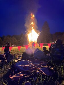Large wooden art structure engulfed in flames at night while festival goers watch from blankets behind red flag barriers