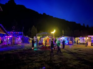 Nighttime festival scene with glowing moon sculpture, illuminated camps, and dramatic canyon silhouette against deep blue sky
