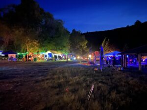Row of illuminated theme camps at night with fire sculpture and colorful lighting against tree line