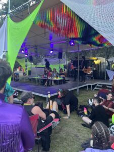 Daytime performance on elevated stage with rainbow streamers overhead, musician with guitar, and audience lounging on grass