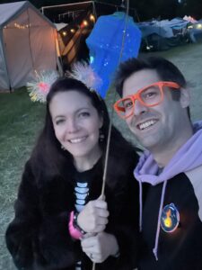 Smiling couple taking selfie at night festival with glowing jellyfish balloon, pom-pom headband, and orange glasses