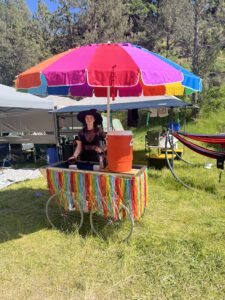 Pickleodeon pickle cart with rainbow ribbon fringe skirt under colorful umbrella, crew member in witch hat serving