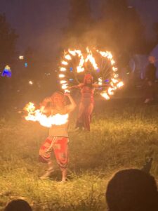 Two fire performers spinning flaming poi and fire fans at night with dramatic flames illuminating the field