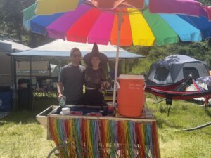 Smiling couple posing behind the Pickleodeon pickle cart with rainbow umbrella and rainbow fringe decoration