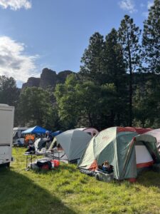 Festival camping area with multiple tents set up on grass with dramatic rocky cliffs and pine trees in background