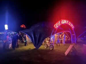 Nighttime festival scene with illuminated archway, colorful lights, and festival-goers gathering