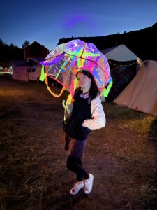 Festival attendee posing with illuminated umbrella decorated with neon glow sticks at dusk