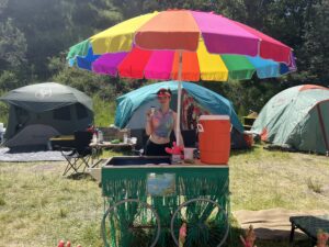 Smiling crew member in tie-dye serving from the decorated Pickleodeon cart on a sunny festival day