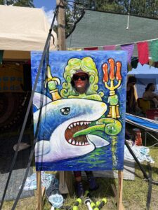 Person posing in painted wooden photo cutout board featuring pickle-themed Poseidon riding a shark