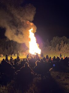 Silhouettes of crowd watching massive bonfire with sparks and smoke rising into night sky at SOAK festival
