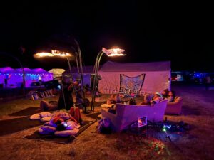 Cozy nighttime lounge area with flame effects overhead, couches, bean bags, and festival goers relaxing under purple lighting