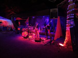 Live band performing on outdoor stage with drums, guitar and bass under dramatic red and blue stage lighting